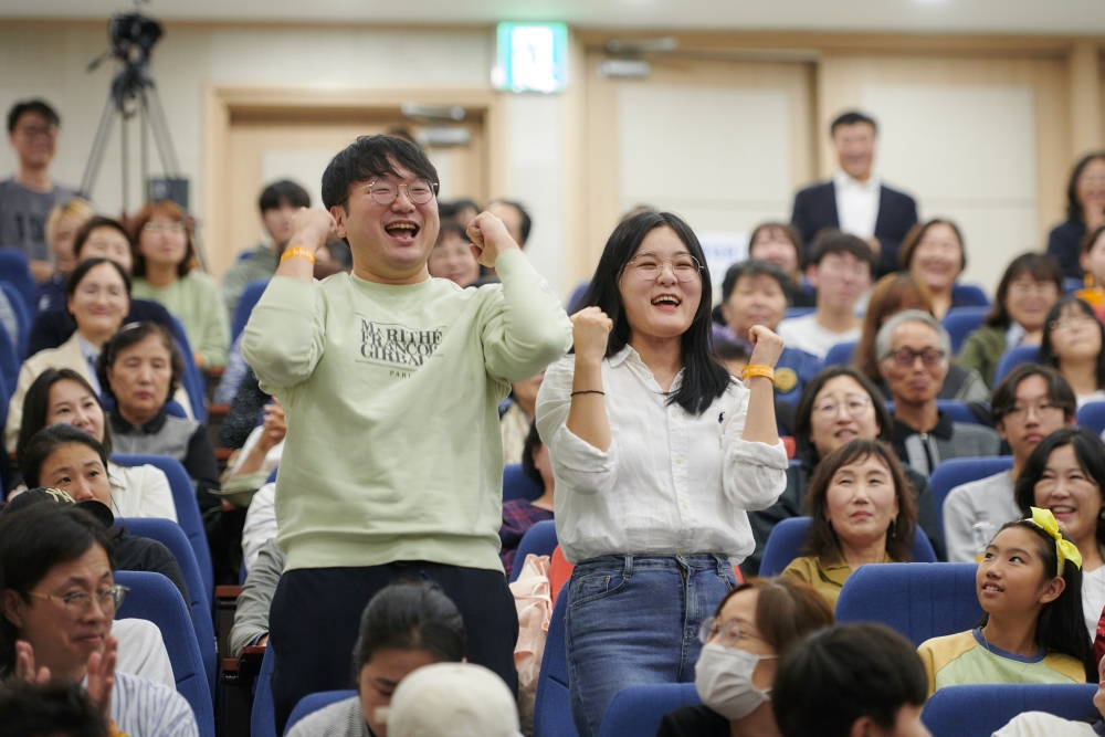 세바시 강연회 객석에서 두 남녀 참석자들이 양손을 불끈쥐며 웃고있는 모습. 주변의 다른 참석자들도 웃으며 이들을 바라보고있다.